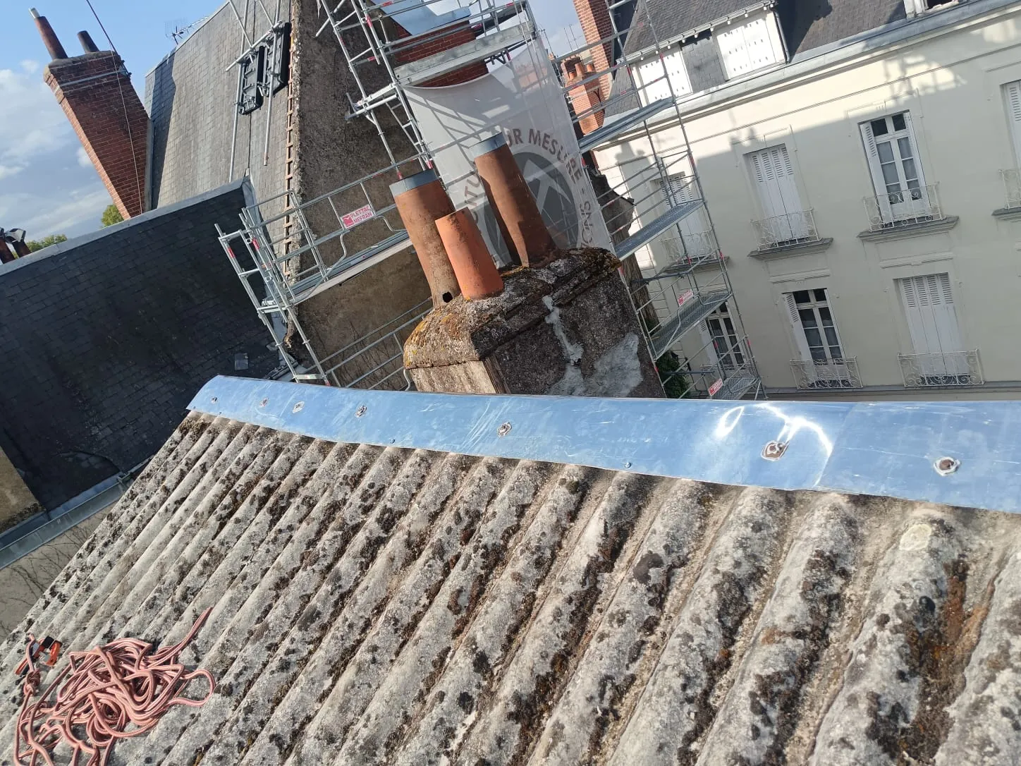 Couvertine en zinc neuve vissée sur un muret de toiture, échafaudage et cheminées de centre ancien derrière