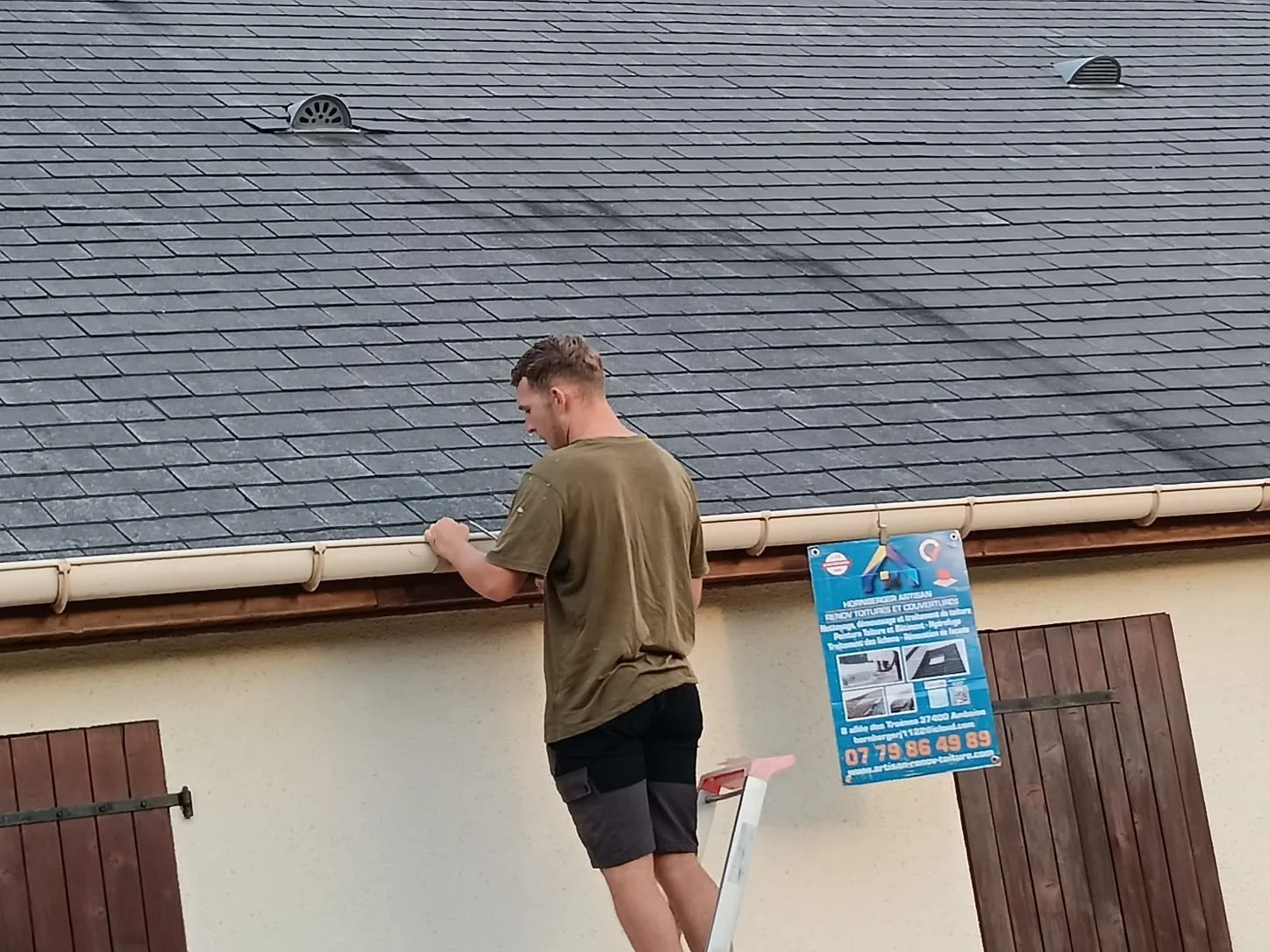 Curage d'une gouttière depuis une échelle le long d'une maison, panneau de chantier posé